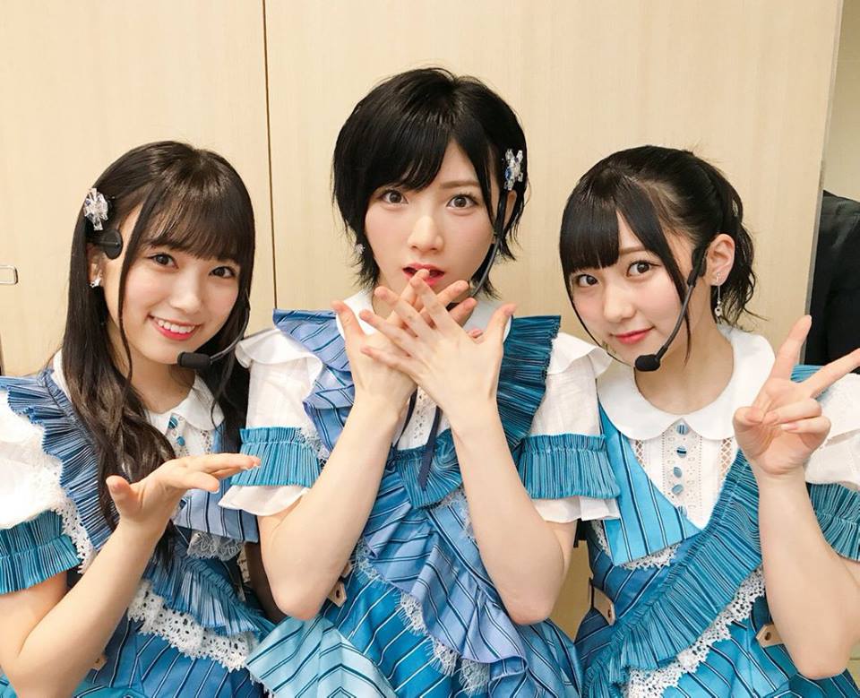 A group of young children sitting at a table - In the image, three young women are the main focus, standing side by side against a beige backdrop. They appear to be dressed in matching blue dresses with white collars, further enhanced by small, decorative hair clips in their hair. The woman in the center of the group, with one hand clasped over her mouth and the other hand raised, seems to be drawing attention or perhaps showing a gesture. The other two women are slightly more composed, with small smiles on their faces. To the right of the frame, the top of another person is visible, but not enough of the person to provide any additional context. The overall scene suggests a staged, celebratory moment, with the women possibly being involved in a performance or promotional event.