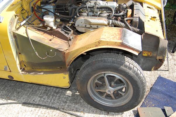 Pistonheads Healey Austin Sprite - The image shows the rear wheel area of a vehicle which appears to be an older-style Volkswagen bus. The wheels are black rubber, and the body parts around them are rusty and corroded, indicative of wear and possible weather elements. We can see various mechanical components and wiring, typical of an open-engine or undercarriage shot. There's also a black tire cover. The overall impression is one of a well-used, potentially vintage vehicle that might require restoration or repair.