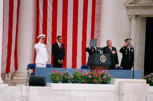 The image captures a formal event, possibly a political ceremony, set against a backdrop of the United States Capitol. Three men, presumably military personnel, are standing behind a rostrum, flanked by American flags on either side. The central figure appears to be speaking at a podium, while the other two soldiers stand at attention. Their professional attire, consisting of military uniforms and ties, contributes to the formal atmosphere of the event. The presence of American flags and the Capitol building further emphasize the significance of the ceremony.