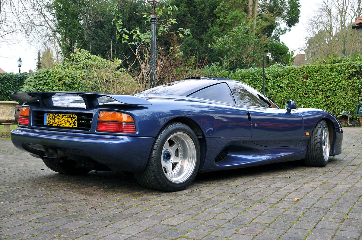 Great Drive in the Jaguar XJR-15 - Page 1 - Jaguar - PistonHeads - The image presents a side view of a vibrant blue sports car, whose aerodynamically sleek design is accentuated by large rear wings, reminiscent of "wing cars" that are often associated with racing and speed. The car is parked on a gravel driveway, its black roof contrasting with the lush greenery of behind it, hinting at a private, possibly suburban setting. The overall scene conveys a sense of tranquility and exclusive vehicular taste.