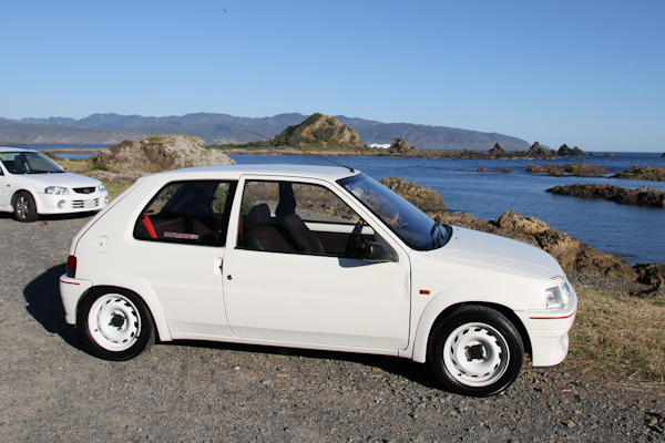 Q for our Peugeot fans - Page 1 - New Zealand - PistonHeads - The image features a white sportscar parked on a gravel surface along a shoreline. A body of water can be seen nearby, with rolling hills and a distant mountain range behind the car. The sky is clear and blue, suggesting a warm, possibly sunny day. In the distance, there appears to be another car parked farther away on the same gravel. The vehicles possess aerodynamic designs with smooth contours and alloy wheels, indicative of performance-oriented features.