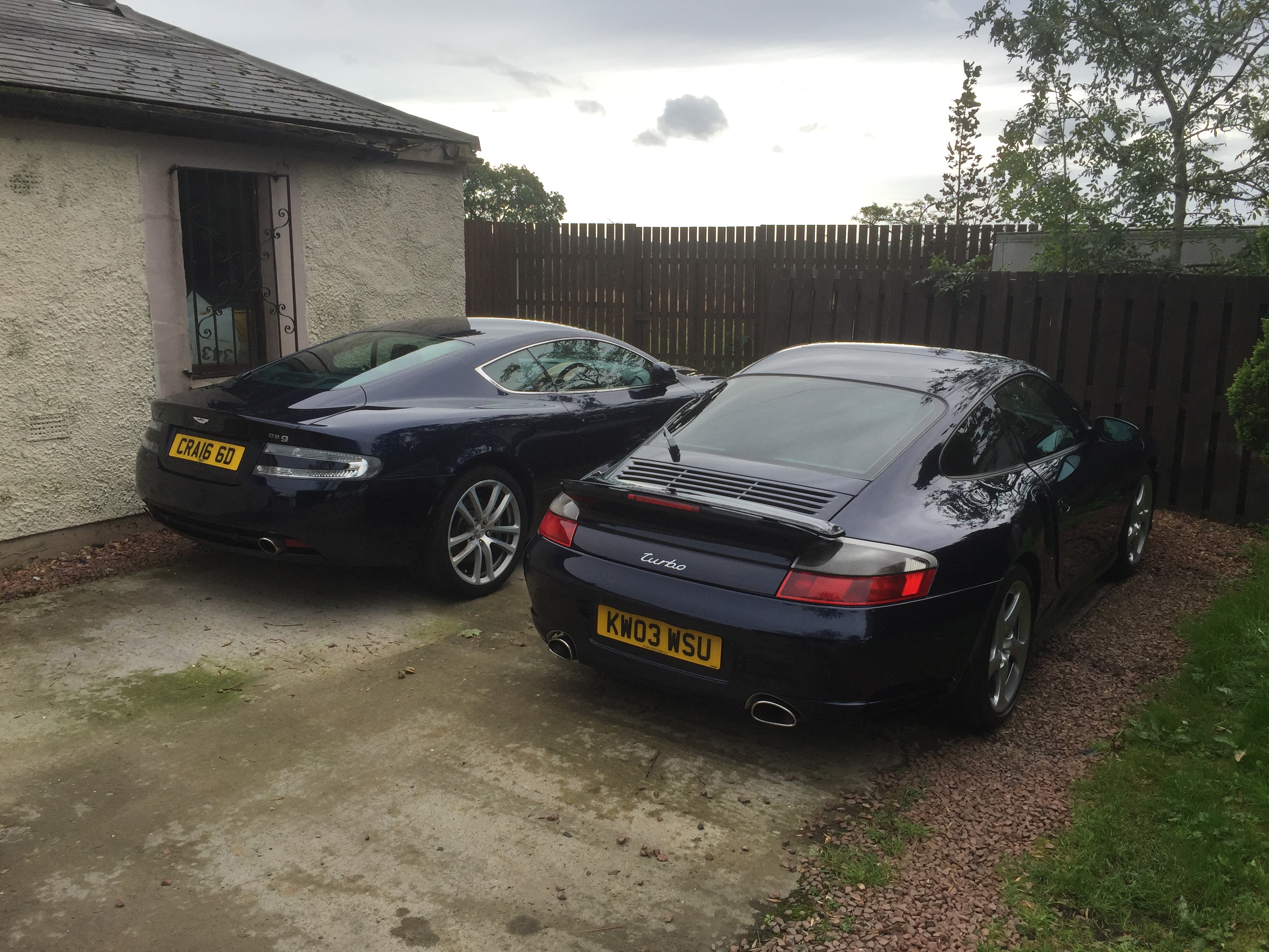 996 gt3 or 996 Turbo. Stressed! - Page 4 - 911/Carrera GT - PistonHeads - The image depicts a tranquil scene featuring two sports cars parked in a gravel lot adjacent to a modest house. The house, partially visible to the left in the image, has a chimney and a sloping roof. The sky background is overcast with a few scattered clouds, suggesting it may be a cool or late evening. The windshields of the cars reflect the ambient light, adding to the serene atmosphere of the setting.