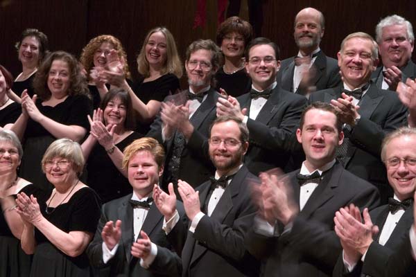 Porsport Pistonheads System Reverse Electric - The image is a black and white photograph of a group of people seated in neat rows, predominantly adults. They are clapping and smiling at the camera, suggesting they are participating in or enjoying a band performance. The individuals are dressed in formal attire, with many men wearing ties, and all people exhibit a cheerful and engaged demeanor. The upper body of someone in the foreground is partially reclined on the person in front of them, indicating interaction or camaraderie among the group. The backdrop is more indistinct, but it appears to be the inside of a concert hall or auditorium.