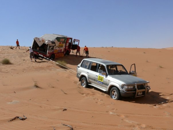 Drives Pistonheads Middle Readers East - This image depicts a robust, white SUV navigating a sandy landscape. The vehicle is partially submerged in the sand, which appears to be kicked up to the side by the tires. The sizable SUV is being tugged uphill by a pair of workers, each pushing from one end. The men are standing in a patch of earth that seems to have a higher water content, contrasting with the surrounding sand. In the background, a large truck tows another car up a hill, adding to the scene of physical labor and the harsh environment.