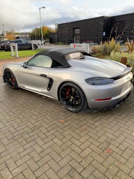 The new 718 Gt4/Spyder are here! - Page 68 - Boxster/Cayman - PistonHeads - The image features a grey Porsche sports car parked outdoors, likely on a driveway. The vehicle is positioned at an angle to the viewer, showcasing its sleek design and distinctive red brake calipers. There's a clear sky above, suggesting it might be a sunny day. In the background, there's a building that appears to be a garage or industrial facility, indicating that this location might be related to automotive production or storage. The overall scene is neatly arranged, with the car being the focal point against the more utilitarian setting of the driveway and building.