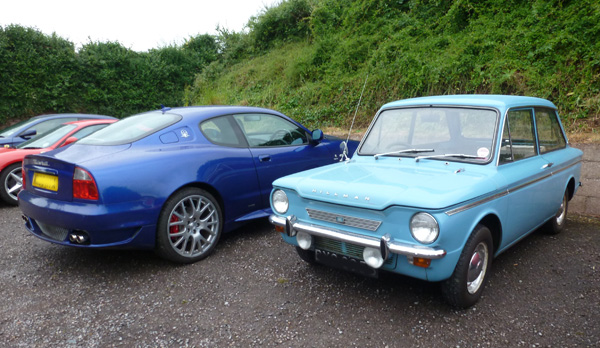 Local meet - Page 1 - South West - PistonHeads - The image shows a small parking area with a focus on three distinctly colored cars. On the left is a sleek, dark blue sports car with a vibrant color and shiny surface, clearly showing the angle of the road. In the center, a small, light blue hatchback is parked, presenting a more classic and less sporty aesthetic. To the right, an older, blue-green vehicle with a vintage appearance adds a contrasting element to the display. Behind these cars, a lush green hillside with foliage provides a serene backdrop to this parking scene.