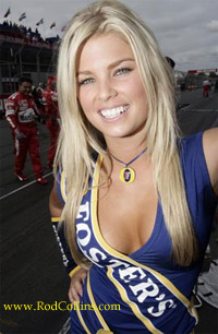 Grid Girls/Pit Babes (Vol 3) - The image is a color photograph featuring a woman smiling at the camera. She is standing with one hand placed on her hip. She has blonde hair and is wearing sleeveless clothing with the word "Racecars" printed in yellow and white across the front. The background of the photo appears to be a racetrack with blurred vehicles and people, indicating that the moment is captured during a race event. There is a watermark on the image with the text "www.RodColin.com."