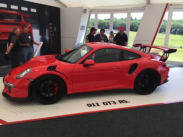 GT2 - Page 111 - Porsche General - PistonHeads - The image shows a vibrant scene featuring a bright red Porsche GT3 RS car as the focal point. The car is placed on a sleek red and white race track-like platform, giving a sense of speed and dynamism. A group of people are gathered around the car, appearing as if they are contemplating or admiring the vehicle. One of the individuals is holding a white lanyard, suggesting they may be part of a formal event or group. The backdrop includes advertisements for automotive parts, adding context to the scene as a possible event or exhibition related to motorsports. The entire setting exudes a sense of anticipation and excitement.