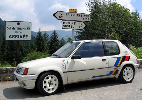 Pistonheads - The image showcases a small, white car parked on the side of a road. The car, adorned with a stripe design on its side, is positioned in front of information signs and near a stone wall. The setting appears to be in a mountainous region. The foliage around and the road itself suggest a rural or semi-rural location. The small size of the car and the multiple numbered signs indicate that this could be a tourist area.