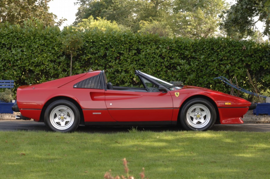 Lets see a picture of your classic(s) - Page 248 - Classic Cars and Yesterday's Heroes - PistonHeads - The image is a photograph of a classic red Ferrari sports car positioned outdoors in what appears to be a well-maintained, grassy area. The car is oriented perpendicular to the viewer, showcasing its front wheel and part of its hood. The vehicle features a convertible top and two front seats. Behind the car, there is a neatly manicured green hedge that borders a paved area, and a blue bench or marker can be seen to the left edge of the image. The sky above is clear, suggesting a bright, warm day.