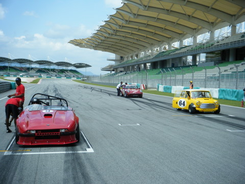 Afos Pistonheads Sepang - The image captures a scene on a race track. Three cars are prominently featured. One is a red car in the foreground, followed by a yellow car in the middle of the track, and a small red car in the background. The red cars have numbers and a description that could be related to the vehicles. The track is near a large structure with a curved roof, which appears to be a grandstand. The lighting suggests it could be daytime. The perspective of the photo helps to create a sense of depth, with the cars and grandstand receding into the distance. The scene conveys the anticipation and excitement commonly associated with motorsport events.