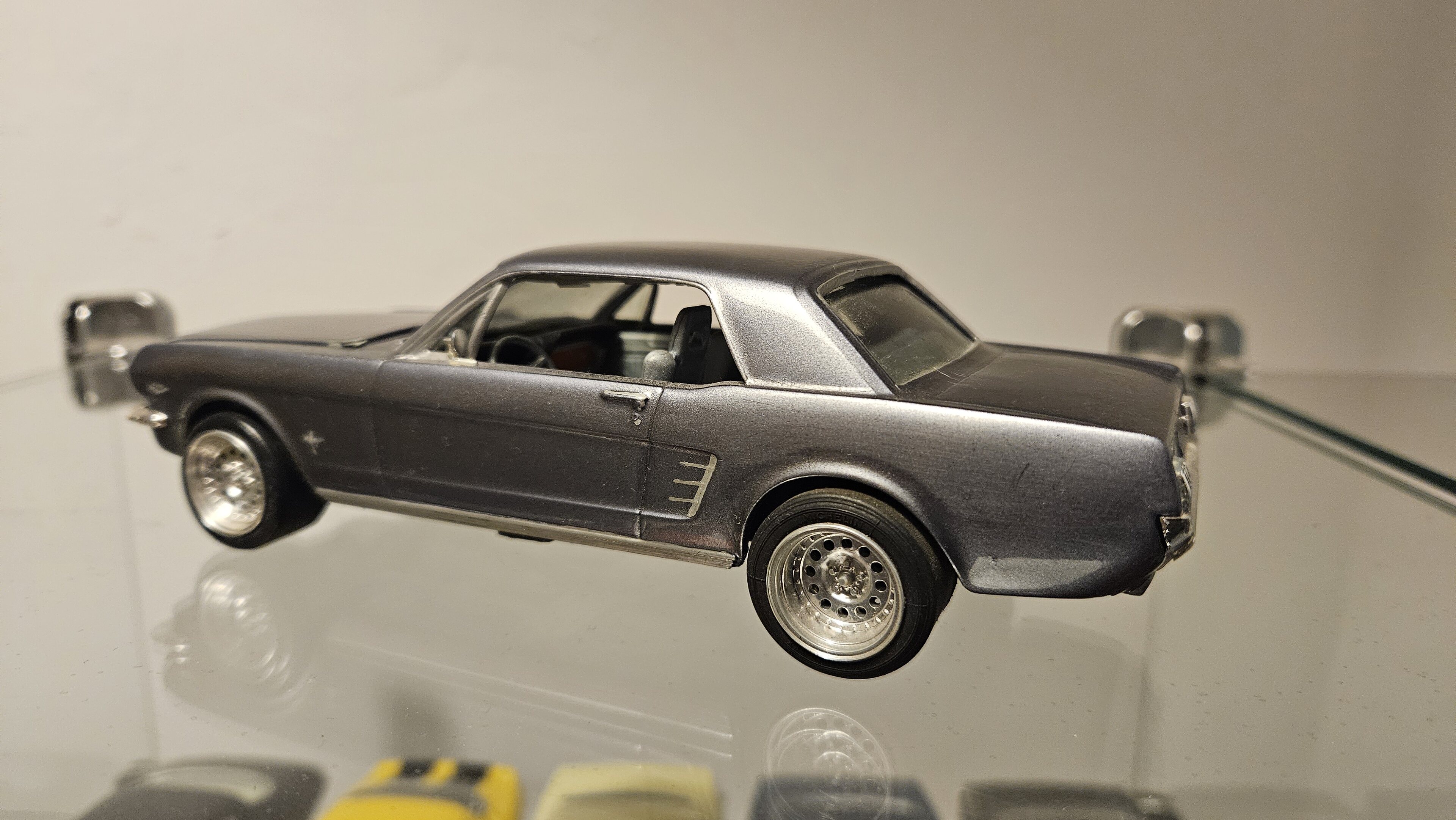 AMT 1:25 1966 Ford Mustang Hardtop - Page 1 - Scale Models - PistonHeads UK - The image shows a detailed model of an old-fashioned, muscle car displayed on a transparent stand. It is situated in a clear case, allowing viewers to appreciate its features from all angles. The car has a shiny silver finish, with the bodywork gleaming. The tires are black and appear to be made of rubber. This miniature automobile is positioned centrally within the image, drawing attention to it as the main subject.