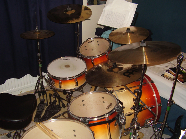 Pistonheads - The image displays a drum set arranged in a typical home setting. The kit appears to be branded "SLK" and features a variety of drums, including a visible tom-tom, bass drum, snare drum, and cymbals. A pair of drumsticks is also seen resting on the base of the hi-hat cymbal. In the background, a single chair is visible, and there is a small amount of text in the upper right corner of the image, possibly indicating the brand or model of the drums.