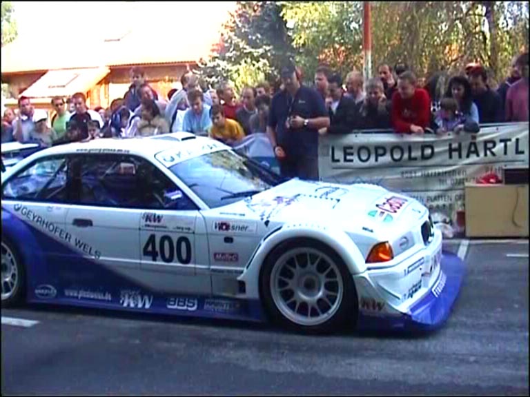 A police car parked on the side of the road - Pistonheads - The image shows a scene on a road where a silver race car with white and blue decals, marked with the number "400" and the name "Leopold Hartl," is prominently displayed. The car is moving or stationary, and the crowd of spectators is gathered on both sides of the road, some standing on the race car. They appear to be watching the car attentively with expressions of excitement or anticipation. The road is lined with fencing, billboards, and signs, and the atmosphere suggests an auto racing event.