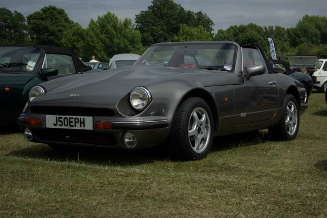 Pistonheads Finished Refurb - The image shows a gray sports car parked on short grass, likely at an outdoor event or gathering. The car has a round license plate that reads "J50EPH" and prominent headlights, suggesting it might be from the 1980s. Its hood is open, displaying the engine, which adds to its sporty appearance. The car is the main subject of the image, and its details make it the center of attention.