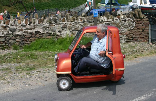 Peel P50 - Page 1 - Classic Cars and Yesterday's Heroes - PistonHeads - The image features an elderly man sitting inside a compact, red, two-wheeled car, which looks like a toy resembling a Smart car. The man is driving the car, and there are no passengers visible. In the background, a stone wall runs along a road with a wooded or greenery backdrop. There doesn't appear to be any text visible in the image. The overall scene is whimsical and charming, emphasizing the humorous and unexpected sight of a small car being driven by a mature individual.