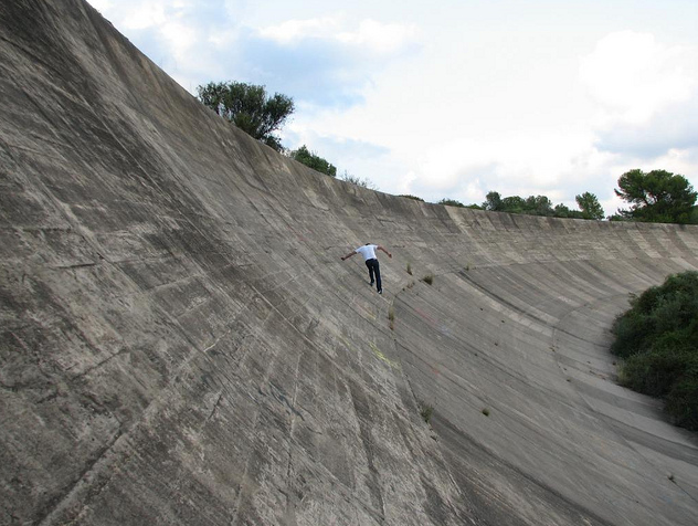 Le Circuit de Reims-Gueux - Page 1 - General Motorsport - PistonHeads - The image showcases a person skateboarding down a steep concrete ramp at a skate park. The skateboarder appears to be in mid-action, riding up the side of the ramp. The park itself is surrounded by a stone wall and features a line of trees in the background. The sky above is filled with low-hanging clouds, suggesting an overcast day. The skateboarder is wearing a white shirt and dark pants.