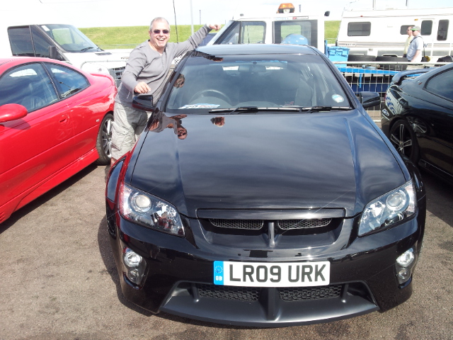 *Forum Event* Performance Vauxhall Show 2012 - Page 9 - HSV & Monaro - PistonHeads - In the image, a man is standing next to a shiny black sports car in a parking lot. The car's doors appear to be open and the hood is closed. There are other vehicles in the background, including a white van and a red car, but they are parked further away from the man and the black car. The man is smiling, suggesting he might be the owner or admirer of the black sports car.