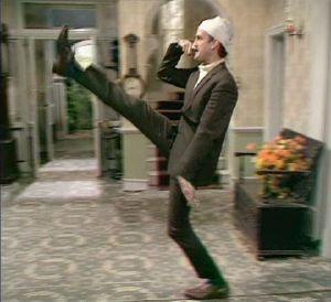 A man standing on a sidewalk talking on a cell phone - Pistonheads - The image shows a man in a formal suit kicking his leg high into the air. His leg is extended straight behind him, with his foot folded under his body. He is also holding a cane in his left hand, which is extended behind him as well. The man is wearing a white turban on his head and is in a room that appears to be a hallway or a transitional space in a home or office. There is a potted plant to his right and a decorative item on the wall in the distance.