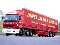 A truck is parked on the side of the road - Pistonheads - The image depicts a red semi-truck with a white cab in the foreground, traveling on a road. The truck is a James Irlam Transport vehicle, identifiable by the "James Irlam and Sons LTD" text on the side of the trailer. The truck carries a flatbed. The cab displays additional branding, with "FLIATE" visible on the windshield. The surroundings include a section of green grass on the side of the road.