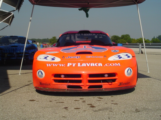 ACR style gen 1 Viper - Page 1 - Vipers - PistonHeads - The image shows an orange racing car with a predominantly orange body. The car is parked outside under a shelter. It features several decals including a large one with the text "PT LAVACA.com" and a smaller one with the number 33. The car has a distinctive front grille and appears ready for a race. The presence of another car in the background suggests that this could be a racing event or a race track.