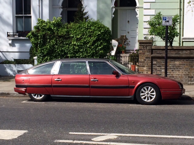 Supercars spotted, some rarities (vol 6) - Page 34 - General Gassing - PistonHeads - The image shows a red, long-wheelbase car parked on the side of a street. It is clear that the car is designed with a rear passenger compartment, as indicated by the two sets of windows with visible door handles. The bodywork of the car appears to be smooth and well-maintained, with a glossy finish. The car is parked in front of a green building with an arched entrance, alongside a narrow walkway and brick wall. There is a parking sign to the right of the vehicle, and the curb suggests a wider parking area. The overall setting appears to be a residential or commercial area.