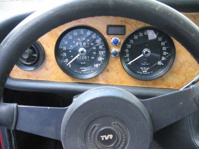 Early TVR Pictures - Page 80 - Classics - PistonHeads - The image captures the driving perspective of a cockpit dashboard. The dashboard itself is a warm brown wood, giving off an air of sophistication. The instruments are clearly visible and include a speedometer, tachometer, and two smaller gauges, possibly for fuel level and engine temperature. A set of steering wheel controls can be seen to the left of the speedometer, suggesting an array of functionalities at the driver's fingertips. The center of the wheel houses what appears to be the brand logo of TRV, indicating the make of the vehicle.