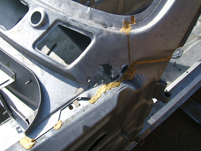 A close up of a car parked on a street - Pistonheads - The image shows a close-up view of a damaged car bumper. The bumper appears to be made of plastic or a similar material, and it is noticeably scrapped and chipped in several areas. There is a large golden-yellow spot that stands out against the grey plastic, which could be rust or damage from another object. The impact of the damage is visible, as the material is torn in places, exposing layers underneath. The background is out of focus, but it appears to be a garage or workshop setting, suggesting that the bumper might have been brought there for repair or replacement.