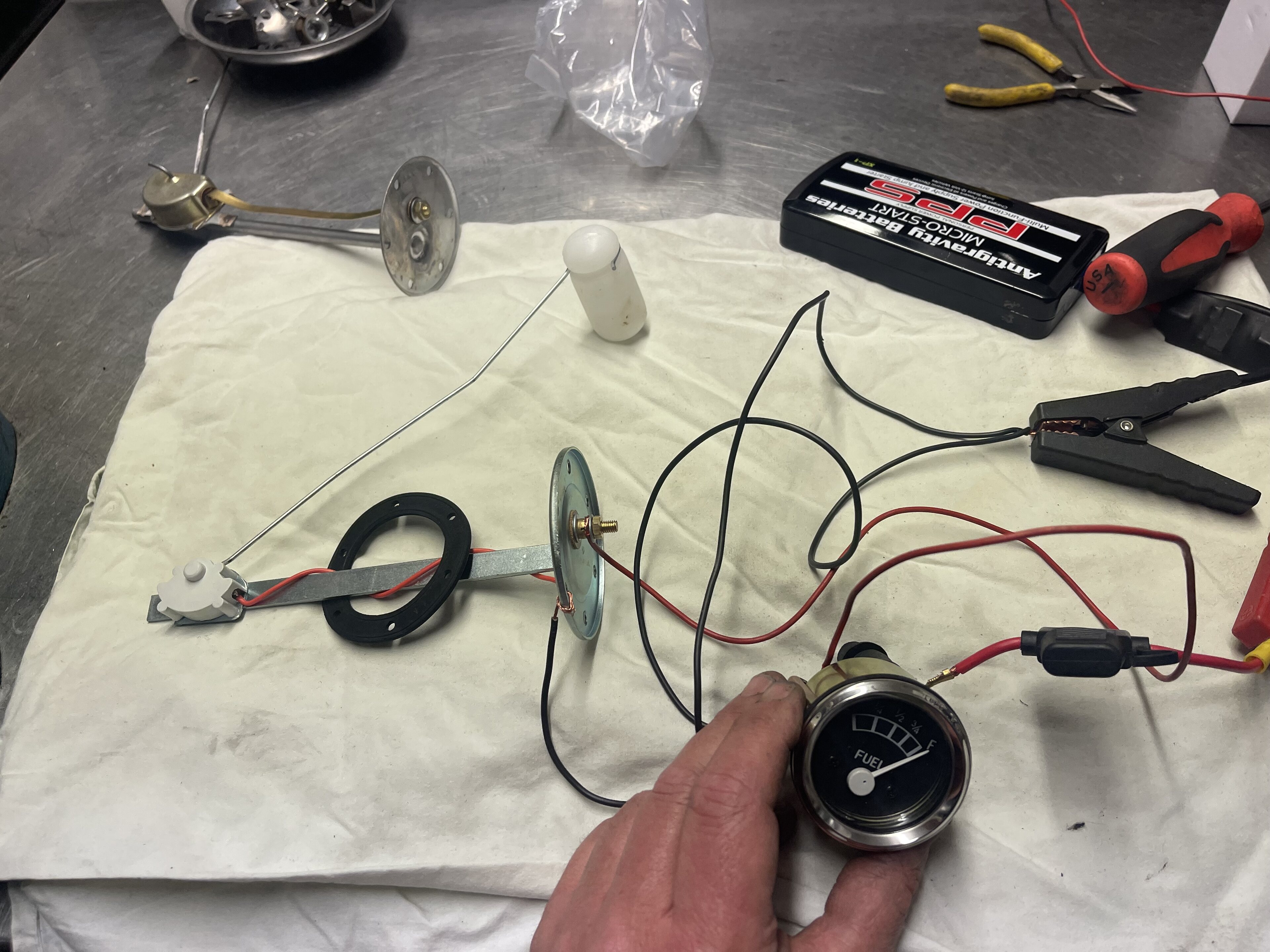 Pistonheads - The image shows a person working with electronic equipment. Various components, including wires, batteries, and possibly a circuit board or other electronic parts, are scattered around on the table. A multimeter is being used to measure something, as indicated by the probes connected to the device. In the background, there is a small amount of clutter that suggests an active workspace. The person's hands indicate they are in the process of setting up or adjusting the equipment.