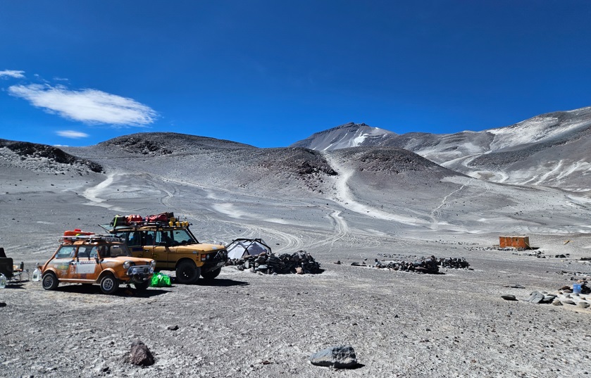 Pistonheads - The image is a panoramic photograph featuring a rugged terrain. In the foreground, there are several vehicles including cars and a truck parked on a dirt road. The sky is clear and blue, suggesting fair weather conditions. Higher up in the background, there is a snow-covered mountain range that appears to be quite remote and likely a popular destination for outdoor enthusiasts. There is a notable lack of greenery, indicating arid or semi-arid conditions. A few scattered rocks can also be seen in the lower left corner of the image. The perspective is from a low angle looking up towards the mountains, which gives a sense of the vastness and grandeur of the natural landscape.