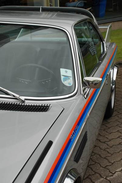 Pistonheads Sunday Service - The image shows a classic car parked outside during the day. The car, specifically a Mustang, is painted in its iconic colors of silver and garnet with blue and orange stripes. The windshield is open, revealing a note inside the vehicle's base. The wheel is on the left side, indicative of a right-hand drive configuration. The Mustang is parked on a paved surface, with a blurred background suggesting a shallow depth of field.