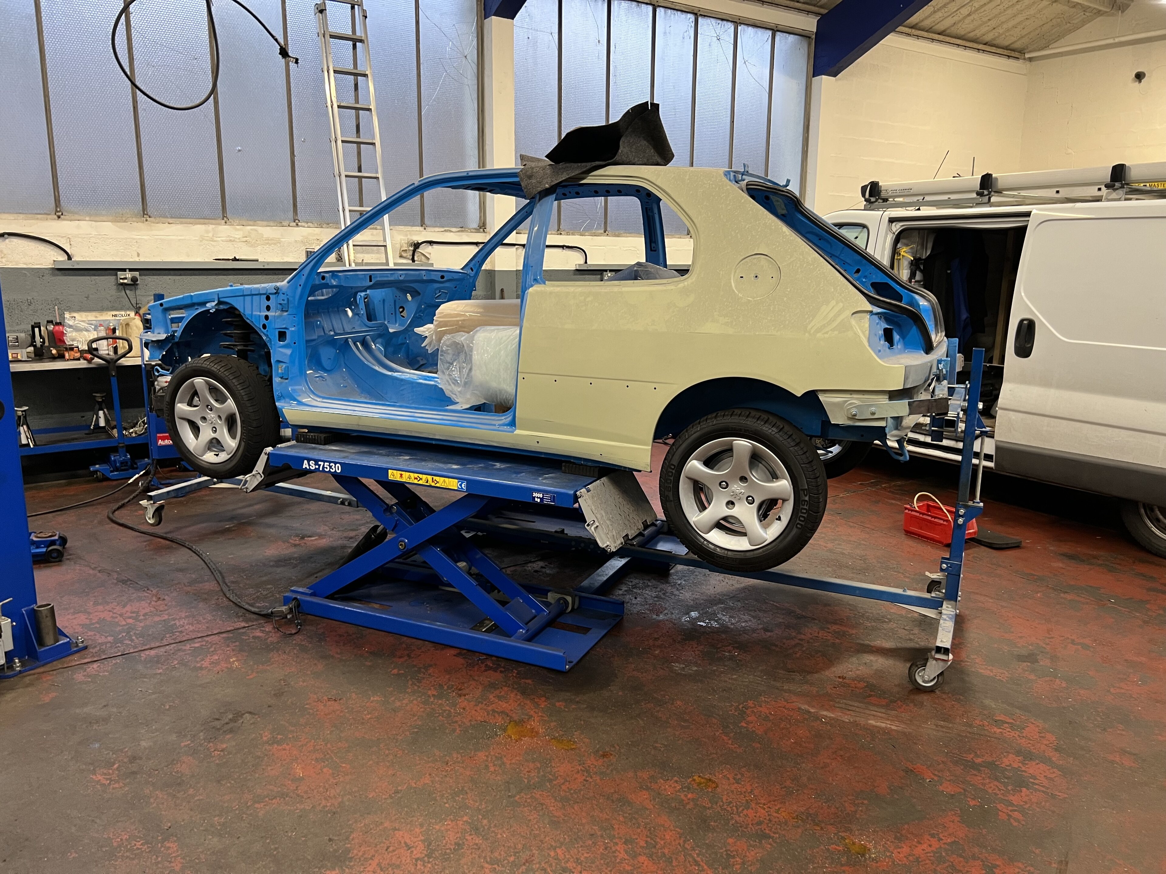 306 GTI-6 - Page 6 - Readers' Cars - PistonHeads UK - The image displays a car body sitting on a hydraulic lift in what appears to be an auto repair or paint shop. The car is positioned for sanding, which has been done in certain areas as evidenced by the rough texture of those spots. The vehicle is an older model hatchback with a distinctive tan interior and a blue exterior that has been stripped down to bare metal. It's mounted on a lift, likely for further work or painting. The background suggests an industrial setting with other vehicles visible in the distance, indicating this might be a professional workshop specializing in car body restoration.