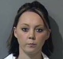 The image depicts the profile of a woman against a neutral background commonly used in law enforcement for mug shots. The woman has light skin and dark hair styled in an untidy updo. She exhibits a neutral expression, looking directly at the camera with a slight frown. The wide shot includes her shoulders and upper back, suggesting a full-body arrest photograph.