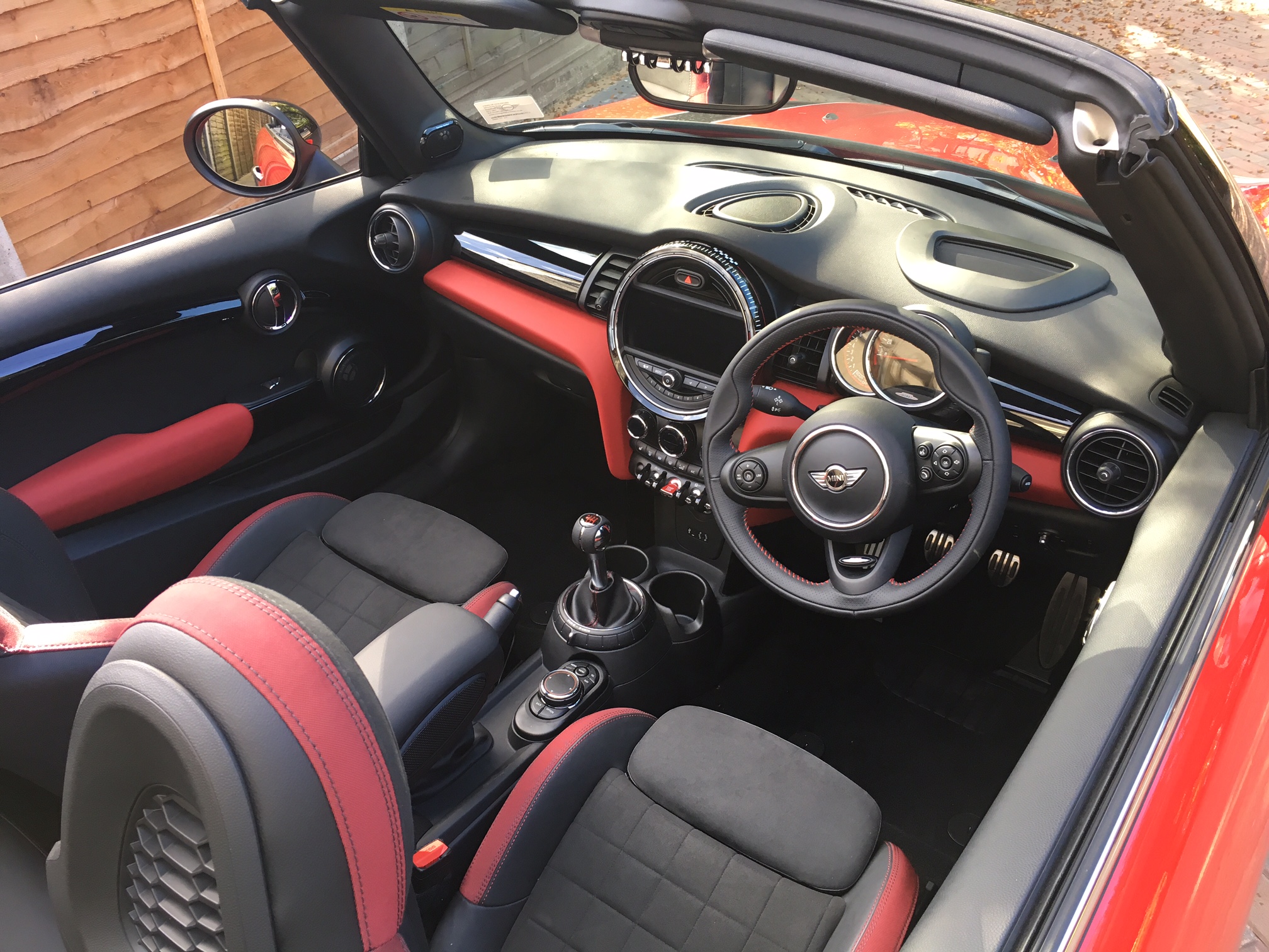 New JCW buyers  - thoughts so far ? - Page 2 - New MINIs - PistonHeads - The image displays the interior cabin of a red convertible car. The car features a black top and red interior accents with the front dashboard in a dark color, possibly black. Both front seats have contrast red stitching on their upholstery, which is black in color. The car is fitted with a manual transmission and the steering wheel appears to be the standard Porsche design, suggesting that this is a Porsche model. The sun roof is retracted, providing a clear view of the sky. The steering wheel sits in the center of the dashboard, with the gear shifter within easy reach of the driver. The seats are head-rested and the driver's seat is positioned towards the center console.