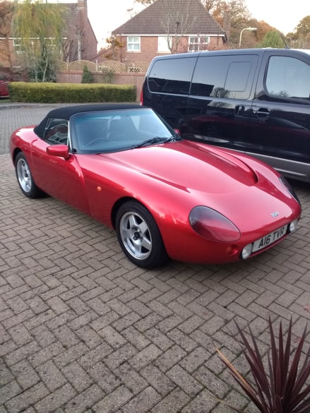 Production start and VIN numbers - Page 3 - Griffith - PistonHeads - The image depicts a vibrant red sports car parked on a driveway. The car is an older model, identifiable by its unique design and the lack of modern features. It's positioned slightly off-center, with its front facing towards the right side of the frame. Behind the car, there's a glimpse of a suburban setting with houses and trees in the background. A person can be seen at the top left corner of the photo, though they are not the main focus of the image. The overall scene suggests a quiet residential area during daylight hours.