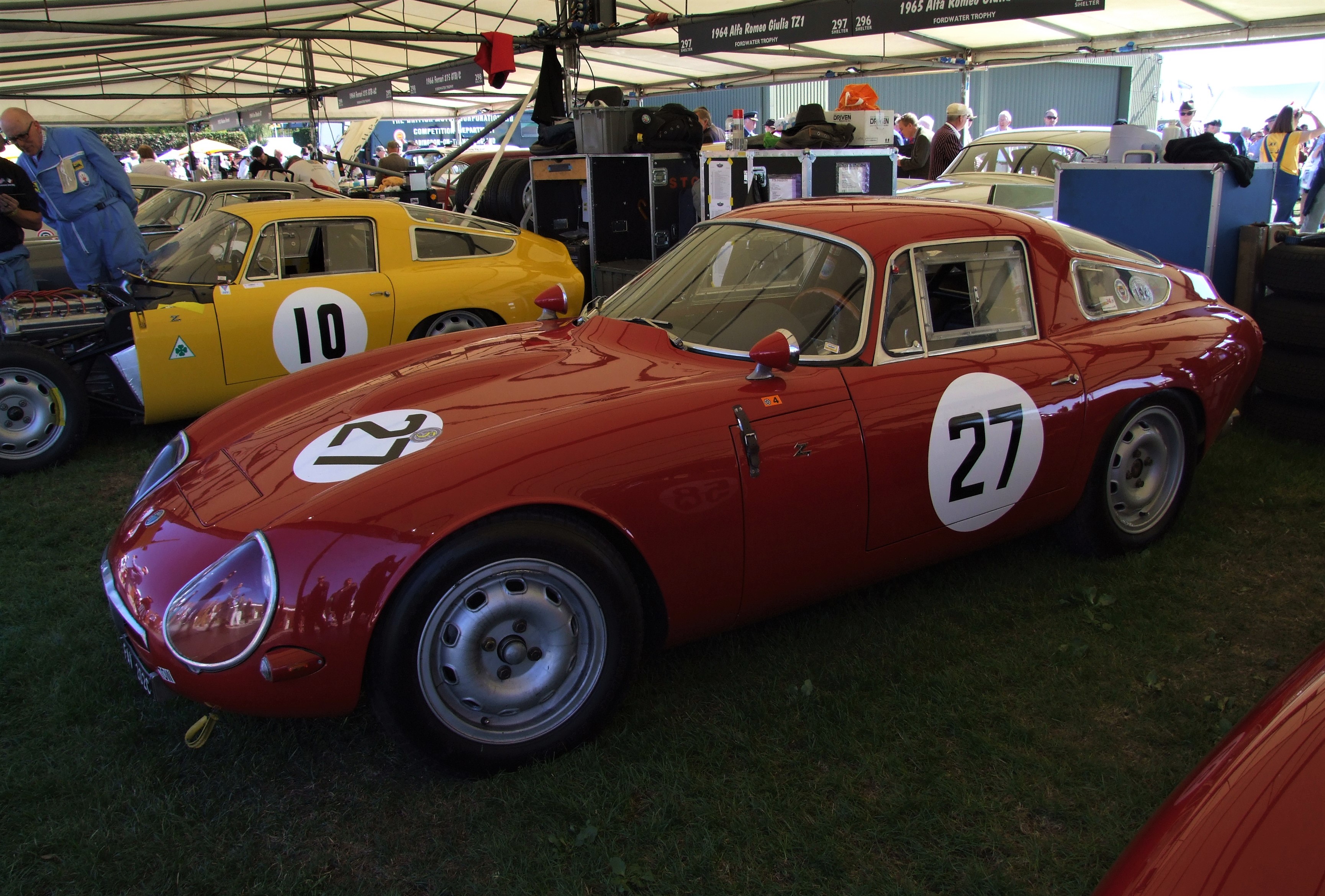 Reflections on this year’s Revival.  - Page 1 - Goodwood Events - PistonHeads - The image showcases a classic car exhibition. There are three classic cars on display, each with its own unique features. One of the cars is a vibrant red race car, adorned with white and black stripes. Adjacent to it, there's another race car in hues of blue and white. The third car, displayed next to the other two, is painted yellow and sports black and white stripes. These cars are parked under a tent, providing shelter from the elements. The scene is lively with several people milling about, admiring the vehicles.