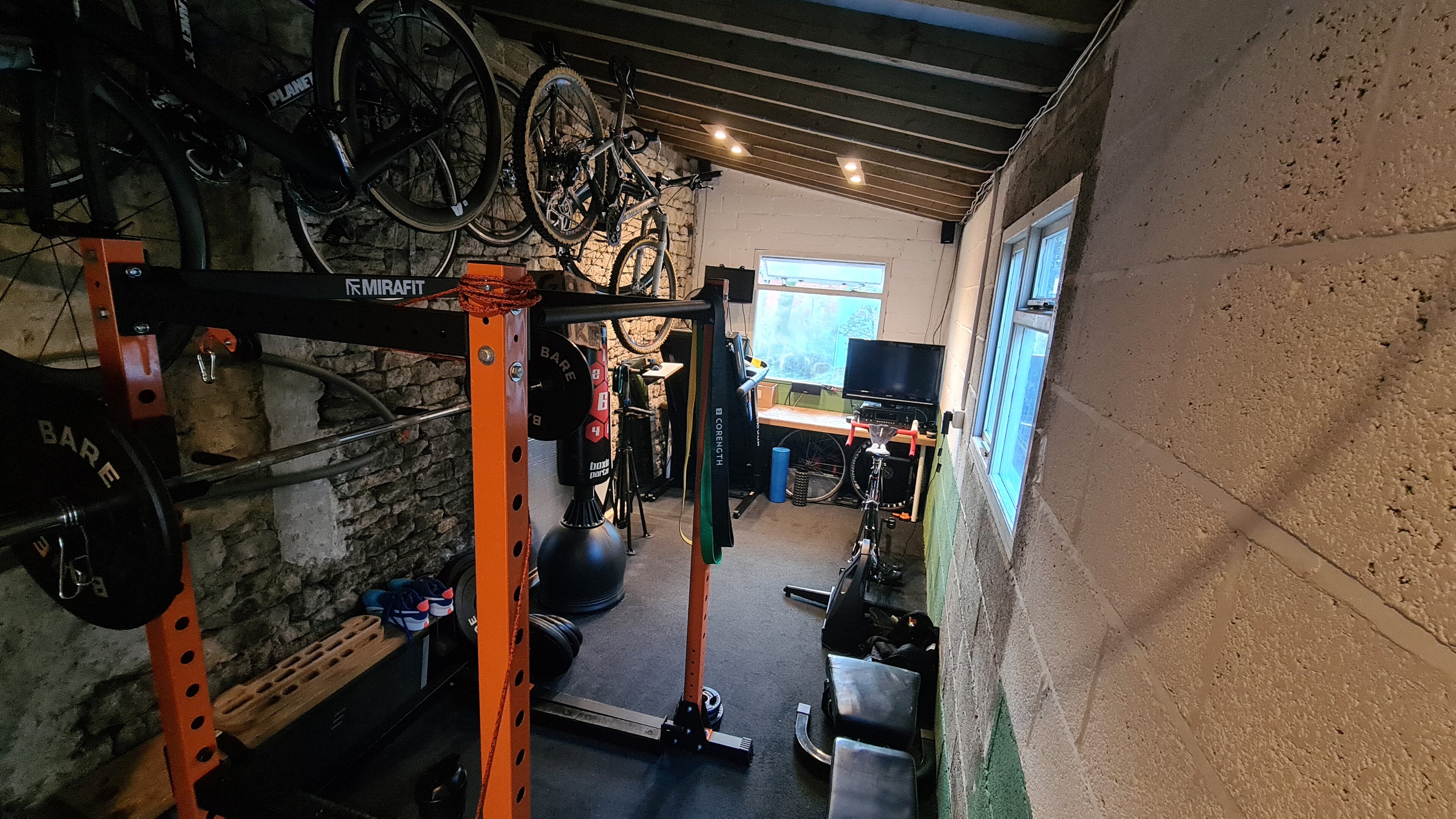 Pistonheads - The image shows a gym setting, likely for weightlifting or CrossFit. There is equipment visible, including several Olympic rings and dumbbells. A bicycle hangs from the ceiling, indicating a possible bike parking area within the gym. The room has a rustic feel with stone walls, and there's a window in one of the walls, letting in natural light. The overall atmosphere appears to be well-lit and organized for physical fitness activities.