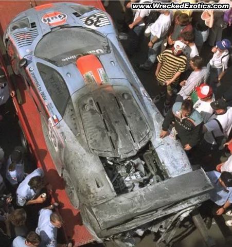 Mclaren Pistonheads Flemke - The image shows a severely damaged race car on a flatbed tractor. The car is stripped of its paint and bodywork, revealing the bare metal beneath. Several individuals are gathered around the car, potentially inspecting or working on it. The car is on a track, and there are various types of equipment scattered around the area, suggesting some form of emergency response or recovery operation.