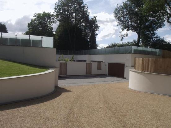 A large pool with a large pool of water - Pistonheads - The image depicts a modern residential property. The house is characterized by its white exterior and a flat roof with an overhanging eave, which adds a unique architectural element. There is a visible wooden deck surrounding the house, blending seamlessly into the landscaped yard. In front of the house, a curved driveway made of gravel serves as a path leading to a closed garage door. The surrounding area consists of a well-manicured lawn dotted with trees. The property appears to be quiet and private, with no visible people or vehicles.