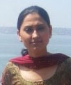 The image is a close-up portrait of a person. The individual appears to be a young woman with dark hair, dressed in what seems to be traditional South Asian attire, possibly a red shawl over a printed top or dress. She is facing the camera directly, displaying a neutral expression. In the background, beyond the person, there is a calming body of water, suggesting a serene outdoor setting. The lighting in the photo is bright, indicating that it was taken during the day in clear weather conditions.