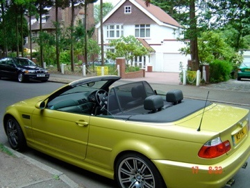 E46 M3 Daily driver? - Page 2 - M Power - PistonHeads - The image shows a vibrant yellow sports car parked on a curved street. The car is convertible with the back roof open, revealing the interior. In the background, there's a residential building with a garage, and the street is lined with trees and residents' houses. There are also other parked cars indicating a residential area. The scene suggests a peaceful suburban setting with a hint of luxury and sportiness.