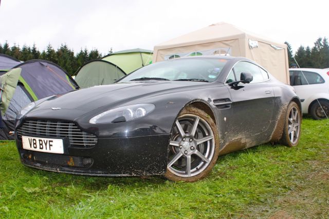 Pistonheads - The image shows a car parked on a grassy surface with mud at its wheels, suggesting recent use on an off-road terrain. The car is a sleek and sporty design, possibly a sports car, characterized by a smooth and aerodynamic body with a sloping hood and side profile. Behind the car, there's a white car, partially visible on the left side of the frame, and a row of colorful tents set up in the background, indicating the presence of an outdoor or camping event. The visual environment, characterized by the mud on the car and the tents, conveys a sense of adventure or a temporary stopover en route to a destination.