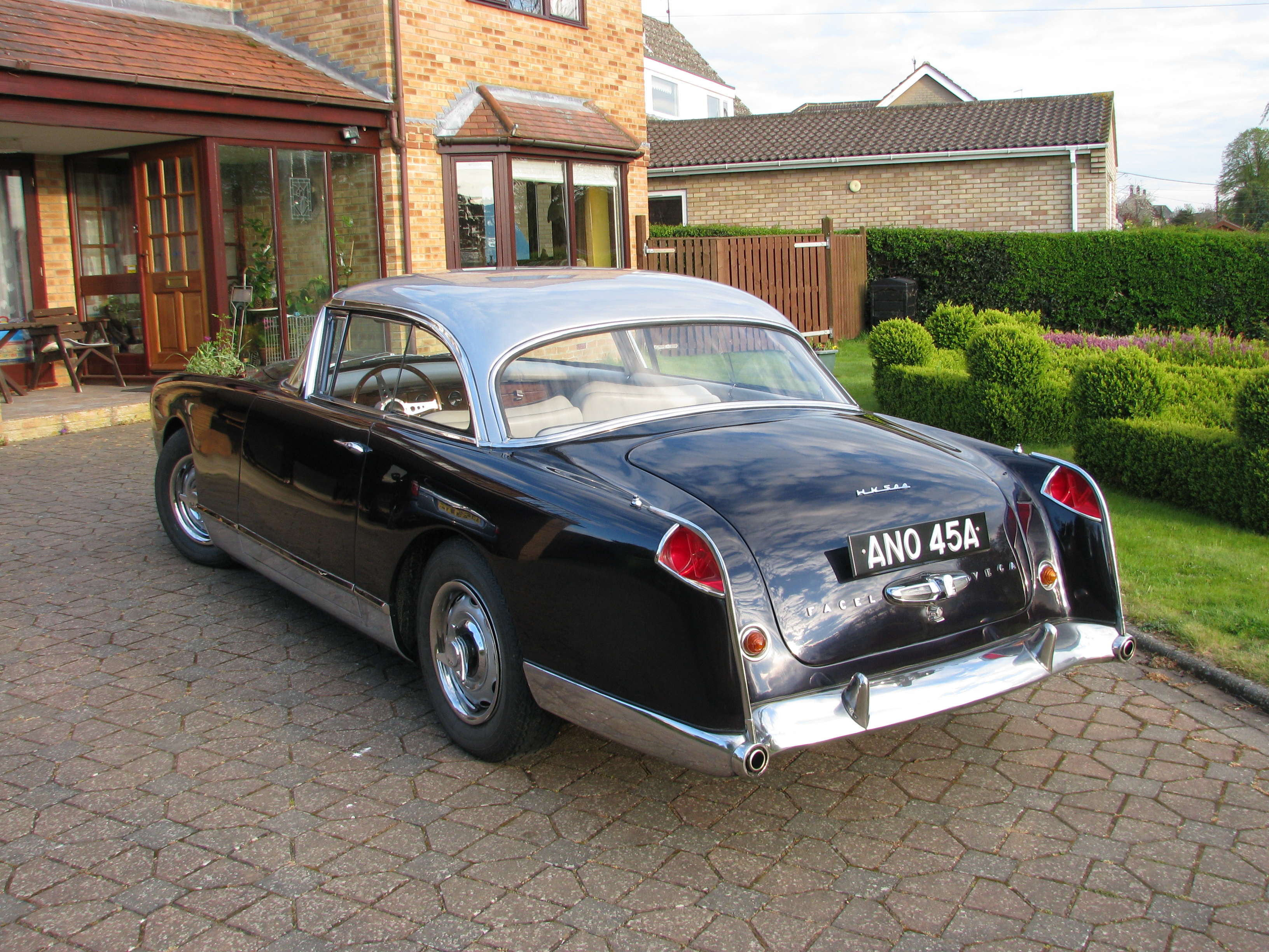 Help Facel Vega, Facel 2 - Page 40 - Classic Cars and Yesterday's Heroes - PistonHeads - The image features a classic Aston Martin parked on a brick driveway in front of a residential house. The car is a two-door model with a retro design, characterized by its hood ornament, round headlights, and distinctive tail lights. The vehicle appears to be in excellent condition, suggesting it might be a collectible or show car. The house is brick with green bushes in the yard, giving the scene a homey and welcoming ambiance. The setting suggests a peaceful suburban area, possibly in the UK given the brand of the car.