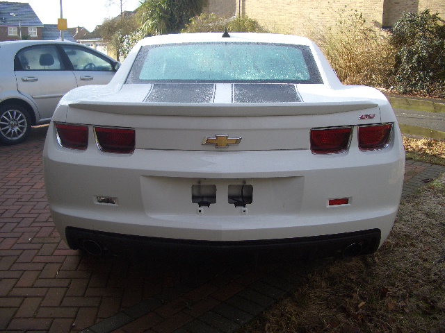 2010 Camaro - Page 1 - Yank Motors - PistonHeads - The image showcases a rear view of a white Chevrolet Corvette located on a brick driveway. The car's taillights are clearly visible and they look pristine. The design of the Corvette, with its aerodynamic rear fender and two round brake lights, is unmistakably sleek and sporty. There is a small black spot on the fender of the car, perhaps a sticker or a mar on the surface. In the background, there's a glimpse of a gray sedan parked parallel to the brick driveway. The overall scene suggests a residential neighborhood with well-maintained vehicles.