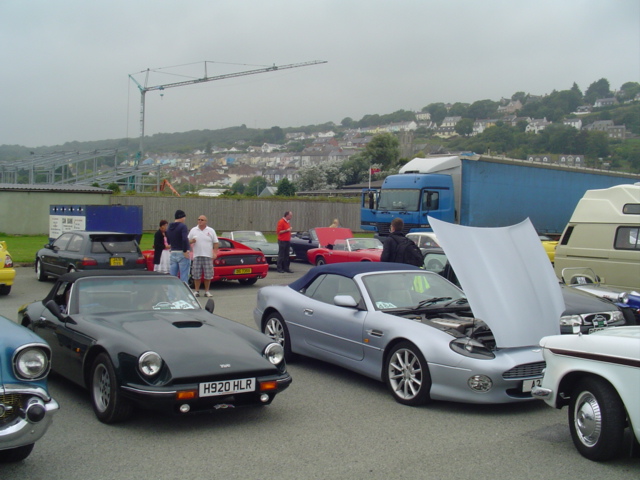 Wales Classic West Pistonheads - This image captures a lively scene at an auto show, where various types of cars and trucks are parked on a tarmac. The setting appears to be a breath of fresh air with a backdrop of rolling hills and trees, creating a scenic contrast to the urban setting of the cars and trucks. There are people milling about in a parking lot, which is filled with an impressive array of vehicles. The attendees seem to be enjoying the exhibition, with one individual taking a closer look at a model car. The image overall exudes the energy and excitement typically associated with such events.