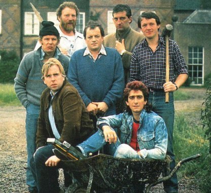 A group of people sitting on a bench - Pistonheads - This image captures a moment on a gravel lot where a group of people, composed of three men standing and one man sitting in a wheelbarrow with a shovel, appear to be engaged in an activity. All individuals are dressed casually, with one man prominently wearing a blue jacket and another in a plaid shirt holding a shovel. The setting suggests a work or outdoor activity, enhanced by the presence of the wheelbarrow and the group's attire. There is no text visible in the image. The photo is shot in a relaxed and candid style, giving the group a casual and laid-back appearance.