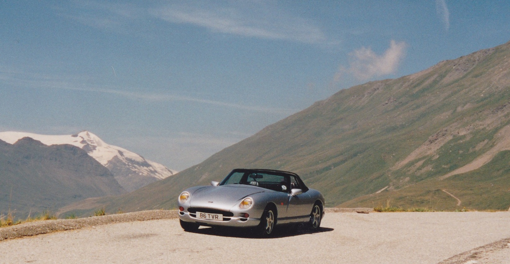 AUGUST PHOTO COMPETITION -  - Page 1 - Photography & Video - PistonHeads UK - The image presents a serene outdoor scene with a silver sports car as the focal point. The car is parked on a dirt road that cuts through the landscape, leading towards snow-capped mountains in the distance. The car's sleek design and shiny surface reflect the sunlight, adding to the overall vibrancy of the image.

The road itself appears well-traveled, suggesting a popular route for tourists or locals alike. The surrounding environment is lush with greenery, indicating that this might be a mountainous area known for its natural beauty. The presence of the mountains in the background adds depth to the scene and creates a sense of vastness.

The composition of the image places the car slightly off-center, drawing attention to the breathtaking scenery around it. Despite the focus on the car, the photographer has effectively managed to capture the essence of the landscape and the tranquility it offers. Overall, the image is a beautiful blend of human engineering and natural splendor.