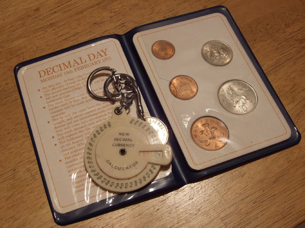 Decimal Day - 40 Years Ago Today - Page 1 - The Lounge - PistonHeads - The image shows an opened pocket watch containing a drawer filled with coins. The drawer has slots for eight coins, and it appears there are four coins in the watch: two one pound coins and two two pound coins. The coins have the image of a character on them, possibly a historical figure or a figure from British culture. There's a note on the watch itself that talks about the history of decimal day on February 18th, possibly explaining the purpose of the coin drawer in the watch. The watch is resting on a wooden surface.