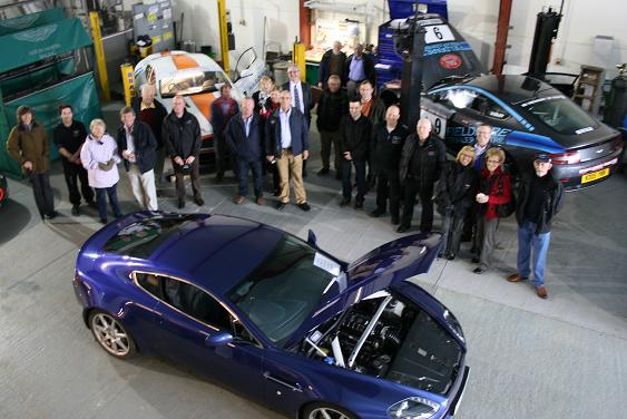 Pistonheads - The image features a group of people gathered around a blue sports car that is open, revealing its engine compartment. The car takes up a significant portion of the image, highlighting its sleek design and inviting onlookers. The hinges of the hood are clearly visible, indicating that the car is well-maintained and possibly on display. Various other cars can be seen in the background, some of which are under a cover and others are fully visible. Lastly, the attire of several people suggests a formal event or expo, where the car is being showcased.