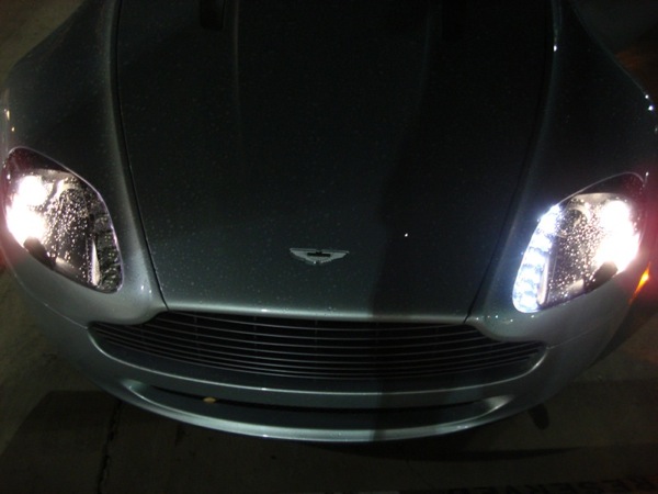 Pistonheads - The image shows a car at night, featuring the front view of the vehicle. It appears to be a solid, dark color, with a front grille visible. The headlights of the car are illuminated, casting a bright light that is reflecting off the wet surface of the ground. The car's design includes a distinctive emblem near the center. The atmosphere suggests that it is a rainy or foggy night, which contributes to the reflection on the ground.